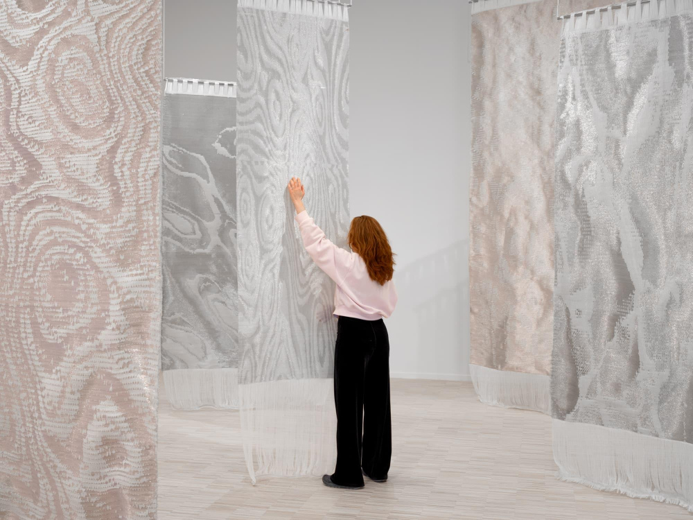 Fabric, Wire, Voice, Water: Weaving Voices as a Merging of MaterialitiesBy Petteri Enroth&nbsp;&nbsp;If I had to pick one phenomenon from the timeline of contemporary art that has heralded and exemplified the significance of materials for artworks, I would probably point toward textile art. With long and varied histories as something homely, crafty, hobby-like and feminine, it entered the halls of fine art institutions slowly and carefully throughout the 20th&nbsp;century. The fabrics made these halls echo with neglected history, homebound activity, nurture, bodily intimacy, unsung labour, cultural chauvinism, and the unfounded principles of high art.&nbsp;Again, during the 21st&nbsp;century, textile art has grown into one of the biggest trends in contemporary art and is therefore bound to find new paths. As things currently stand in the artworld, this is likely to happen through merging with other materials and media.A rather recent and still quite rare development is combining textiles with sound. Pearla Pigao’s&nbsp;Weaving Voices&nbsp;does this in an original way, one that is useful to contextualize a little.*An intertwinement of textiles and sound is not far-fetched. Just as we are almost constantly in touch with fabrics and fibres, there is no point in our lives that would be completely without sound. In what we think of and live through as everydayness, textiles and sound surround us in an intimate, supportive way. They conduct our atmospheres and mark our senses of place and bodily positioning in similar, often soft and flowing ways – in contrast to other everyday materialities like the asphalt we walk on, the plastic keyboards we slam, or the concrete walls that confine and protect us. Fabric and sound share something in their relation to the body. We notice a cold toe like a cessative noise.Touch and hearing, skin and ear, merge already at the beginning of our lives, in the womb. Once hearing starts to develop at around 18 weeks of pregnancy, the two senses are close to indistinguishable, both mediated by water. They continue to bear similarities later on, when we leave water behind and begin our journey surrounded by air. Hearing someone speak, play an instrument, let alone whisper in our ear is quite literally touching through air, vibrating this mixture of gases between us.&nbsp;Weaving Voices&nbsp;builds on this connection.To do this, the work uses modern technology. Incidentally, technology has also become as much an often-unnoticed part of the quotidian flow of our lives as fabrics and sound. I might be standing at traffic lights, listening to music from wireless headphones surrounded by hybrid-powered cars with a high-voltage power line humming above me while being recorded by a CCTV camera and pierced by radio waves. Technology is, as it were, human-sized in the sense of being omnipresent, low-threshold and easy, and often necessary, to take for granted.Weaving Voices&nbsp;incorporates a sophisticated set of audio technology with hundreds of meters of cable, a modular synthesizer, custom-built electronics, software and such. Listed like this, such a setup might evoke a sense of cold, inhuman modernism. Indeed, when sound art established in the 1970’s, it was often wilfully distant from our everyday concerns and scales, mediating scientific concepts and the estranging potential of sound technology – or, in the field of acoustic ecology, tried to harness this technology to re-create the authentic reality of distant soundscapes, i.e. to use technology so meticulously that it would disappear into itself.This DNA of sound art is present in&nbsp;Weaving Voices, but the work also elaborates on a more recent development within it, one that doesn’t shun the human voice, musicality, and bodily interaction. The work addresses the exhibition space not only as physical, aural or atmospheric but also as social and communal. The way I move in and with the work matters on all these levels. By turning movements into sound,&nbsp;Weaving Voices&nbsp;thematizes the simultaneous materiality, sociality and choreographic nature of the body, emphasized by the performance by four contemporary dancers that has been part of the work since its first exhibition at Kunstnernes Hus in Oslo in 2025.In short, the work makes the air and our movements in it “tangible”. I move, a voice answers – and not just any voice, but part of a composed whole. I can unearth it from the hanging fabrics and affect it to an extent, but it has a heart, structure and trajectory of its own, and only parts of it become audible at a given moment.&nbsp;As if we could make the air ripple, surge and wave in a way that we usually associate with water, a material that&nbsp;Weaving Voices&nbsp;indeed incorporates.&nbsp;Ripples in water often have to do with moments of earthing solitude, quiet reflection, and coming down from the heights of our speculations and aims. And so, to touch the water of&nbsp;Weaving Voices, we must kneel down, altar-like, as if to summon. I can’t avoid thinking of Narcissus here, too: we can be reflected in this water, but only as sound and through an existing composition, by breaking the surface and bringing forth something other than ourselves.*However, we do also exist as visible reflections within&nbsp;Weaving Voices, only not through mirrors or lenses but as electronically mediated biomorphic forms on a board of LED lights.&nbsp;The element makes me think of a kind of cultural history of giving sound visible forms, for which we seem to have a fascination, even a pressure. This thread runs from the Biblical burning bush to Ernst Chladni’s famous namesake sand boards to modern acoustics with its reverberation patterns, refraction visualisations and equations. This time, the combination of sound and light sequences resembles something of a “close encounter of the third kind” with a version of ourselves that is as much wired and mediated as it is immediately corporal.&nbsp;To me, all these aspects of&nbsp;Weaving Voices&nbsp;come together as a curious tension between things like estrangement and everydayness, composition and spontaneity, movement and choreography, participation and otherness, site-specific materiality and abstraction. This is exemplified by the voices of the composition that are as clearly human as they are technologically structured, equalling the undeniable meshing of fabric with metal.Through these tensions, the work does not dramatize or “reveal” the gulf between the immediate and the mediated, sensible appearances and invisible machinations, but rather lets us approach the difference in many modes. I can be playfully exorcising notes from the hanging weaves and the water’s surface in child-like fascination and then look at the LED wall and become aware of how it is very hard to do anything in the contemporary world without being visually presented and represented in ways I do not mean or even want.&nbsp;Most of all, I tend to think of&nbsp;Weaving Voices&nbsp;as a reminder that what we call “air” and treat as emptiness between things is in fact never empty. Rather, it is a strange, soothing, ever-flowing layer around our planet against the emptiness of space, and is constantly alive with vibration, sound, atmosphere, meaning and, most recently, signal and current.